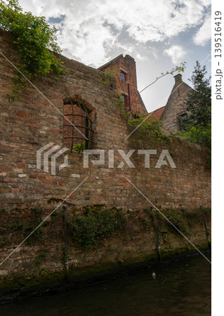 Old brick canal wall in Bruges shows arched opening, ivy, weathered stone base, historic rooftops, quiet water. Heritage tourism, architecture marketing, travel content, urban history 139516419