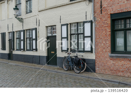 Bicycle with pannier stands beside white brick house facades on cobblestone street in Bruges, Belgium, shuttered windows, muted urban mood. Cycling culture, architecture, sustainable mobility Bicycle with pannier stands beside white brick house facades on cobblestone street in Bruges, Belgium, shuttered windows, muted urban mood. Cycling culture, architecture, sustainable mobility 139516420