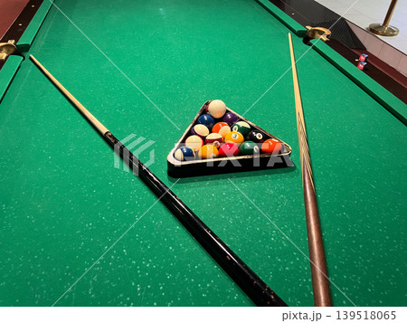 A pool table shows balls in a triangle shape while two cues sit ready for use in a game room during the evening hours A pool table shows balls in a triangle shape while two cues sit ready for use in a game room during the evening hours 139518065