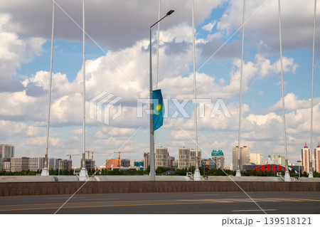 Flag of Kazakhstan hanging on a lamppost on the Arkar bridge. Urban landscape with city buildings and a cloudy sky in the background. Patriotic symbol in Astana, national holiday concept. 139518121