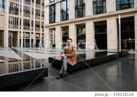 Man sitting on a stone bench in a modern office courtyard. Businessman resting during a break near a city building. Urban lifestyle and corporate architecture in a professional setting. 139518128