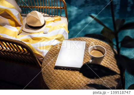 Cozy outdoor setting featuring a wicker chair with a yellow striped blanket, a sun hat, a blank notebook, and a cup of coffee beside a swimming pool 139518747