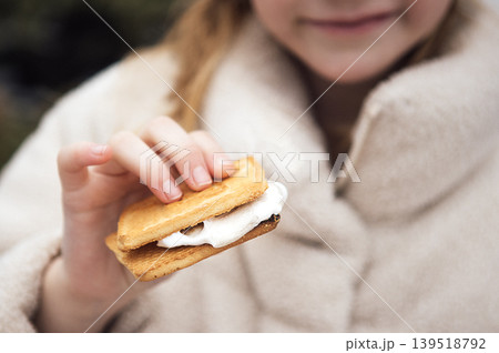 Child holding a smore dessert made of graham crackers, marshmallow, and chocolate, wearing a cozy beige coat, with a blurred outdoor background Child holding a smore dessert made of graham crackers, marshmallow, and chocolate, wearing a cozy beige coat, with a blurred outdoor background 139518792