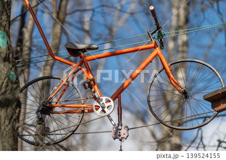 Vibrant bike adventure. Suspended bicycle among trees. Bike hanging over forest with safety equipment. Colorful bicycle sways above woodland with protective ropes 139524155