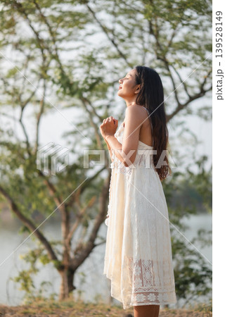 Elegant woman in white lace dress standing beside lakeside trees with eyes closed and hands clasped, enjoying warm sunlight, calm nature, mindful breathing, summer peace, and serene outdoor lifestyle. 139528149
