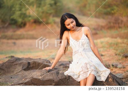 Woman in white lace dress sitting on sunlit rocks with gentle smile, warm outdoor nature portrait, bohemian summer fashion, peaceful lifestyle, wellness, relaxation, travel, and soft rustic beauty. Woman in white lace dress sitting on sunlit rocks with gentle smile, warm outdoor nature portrait, bohemian summer fashion, peaceful lifestyle, wellness, relaxation, travel, and soft rustic beauty. 139528158