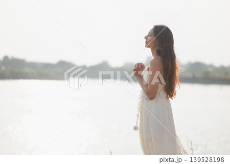 Elegant woman in white lace dress standing beside bright lake in profile with clasped hands, feeling calm sunlight, mindful gratitude, peaceful summer travel, nature wellness, and serenity. Elegant woman in white lace dress standing beside bright lake in profile with clasped hands, feeling calm sunlight, mindful gratitude, peaceful summer travel, nature wellness, and serenity. 139528198