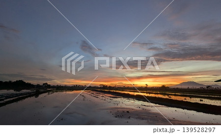 A stunning tropical sunrise casts warm light over flooded paddy fields, reflections mirroring the vibrant sky. Silhouettes of trees, a distant mountain create a peaceful, rustic scene. A stunning tropical sunrise casts warm light over flooded paddy fields, reflections mirroring the vibrant sky. Silhouettes of trees, a distant mountain create a peaceful, rustic scene. 139528297