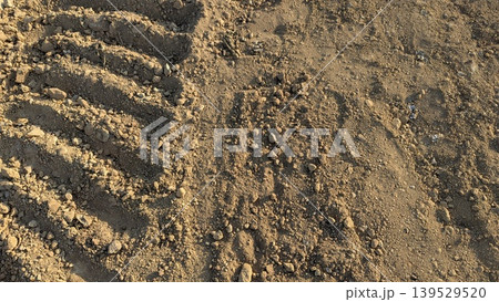 Dry brown soil features rough textures, tractor tire tracks, scattered stones. This natural earth background captures agricultural land under warm sunlight. Perfect for farming, construction themes. 139529520