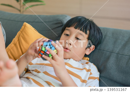 Smart Asian kid lying on a comfortable gray sofa, concentrating on solving a colorful dodecahedron puzzle toy. Child brain development, logical thinking, education and creative leisure time at home. 139531167