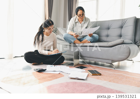 Two women manage household budget at home with reports, smartphone, laptop, and notes, showing financial planning, expense tracking and teamwork in a bright modern living room. Two women manage household budget at home with reports, smartphone, laptop, and notes, showing financial planning, expense tracking and teamwork in a bright modern living room. 139531321
