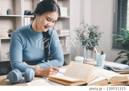 Young Asian woman writing study notes at home desk with open books, coffee and headphones nearby. Focused self study, exam preparation, reading, education and productivity concept. Young Asian woman writing study notes at home desk with open books, coffee and headphones nearby. Focused self study, exam preparation, reading, education and productivity concept. 139531325