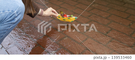 A person placing a yellow origami boat decorated with green clouds into a puddle on a red brick sidewalk A person placing a yellow origami boat decorated with green clouds into a puddle on a red brick sidewalk 139533460