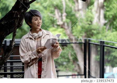 elder 50+ woman stand relax read book outdoor green tree elder 50+ woman stand relax read book outdoor green tree 139537473