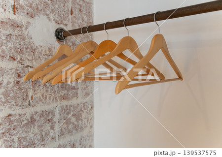 Wooden clothes hangers hang on metal rail against white wall, exposed brick, creating minimal interior detail with clean boutique mood. Retail design, closet organization, home staging, hospitality 139537575