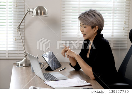 Elder Business woman is sitting at a desk with a laptop and a tablet 139537780