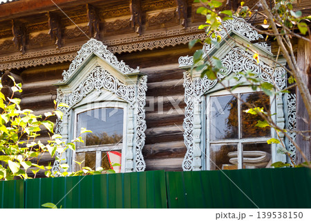 Intricately carved windows adorn an old wooden log house, showcasing traditional Russian craftsmanship and heritage, partially obscured by lush green foliage 139538150