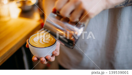 Cappuccino preparation. Hot beverage. Man hand pouring steamed milk from steel jug in espresso coffee mug making white foam pattern. Cappuccino preparation. Hot beverage. Man hand pouring steamed milk from steel jug in espresso coffee mug making white foam pattern. 139539001