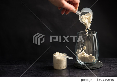 Protein drink. Hand pouring protein powder in a drinking glass. Dark background. Body building, muscle growth and food supplement concept 139539640