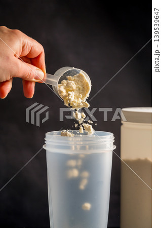 Protein drink. Hand pouring protein powder in a white shaker. Plastic tube on dark background. Body building, muscle growth and food supplement concept 139539647