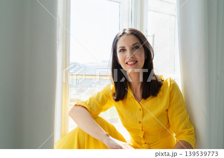 A Cheerful Woman Dressed in a Vibrant Yellow Dress is Posing Gracefully and Happily Near the Bright Window A Cheerful Woman Dressed in a Vibrant Yellow Dress is Posing Gracefully and Happily Near the Bright Window 139539773