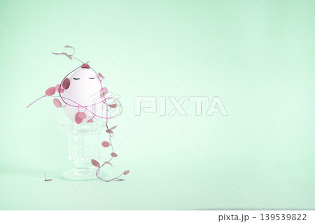 Decorative pink plant with smiling face sits on a glass pedestal surrounded by trailing vines against a soft mint green background 139539822
