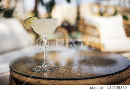 Margarita cocktail in a clear glass with lime wedge on a woven table, blurred background of outdoor seating area with rattan furniture and greenery Margarita cocktail in a clear glass with lime wedge on a woven table, blurred background of outdoor seating area with rattan furniture and greenery 139539830