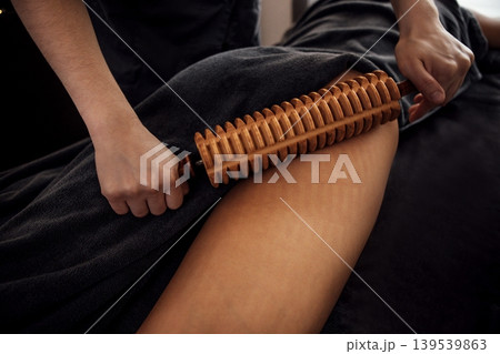 Female massage therapist using wooden roller on client's thigh, demonstrating therapeutic technique in a serene spa environment with soft lighting and calming atmosphere 139539863