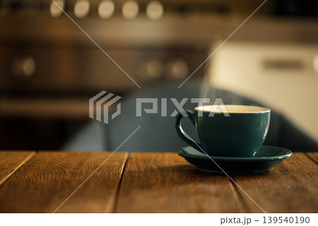 Hot steaming cup of coffee in a teal ceramic mug on a wooden table with a blurred kitchen background featuring metallic appliances and warm lighting Hot steaming cup of coffee in a teal ceramic mug on a wooden table with a blurred kitchen background featuring metallic appliances and warm lighting 139540190