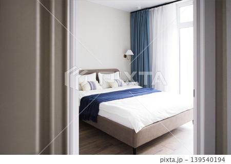 Modern bedroom interior featuring a neatly made bed with white linens and blue throw blanket, large windows with sheer curtains, and wooden flooring visible through an open door 139540194