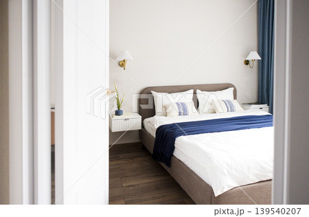Modern bedroom interior featuring a large bed with white linens and blue throw blanket, bedside lamps, and decorative plant in a stylish, minimalist design Modern bedroom interior featuring a large bed with white linens and blue throw blanket, bedside lamps, and decorative plant in a stylish, minimalist design 139540207