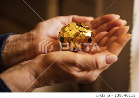 Hands holding a decorative golden egg covered in shiny coins, showcasing intricate details and textures, with a soft background enhancing the visual appeal of the object Hands holding a decorative golden egg covered in shiny coins, showcasing intricate details and textures, with a soft background enhancing the visual appeal of the object 139540218