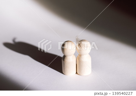 Two wooden figurines standing close together casting shadows on a white surface, highlighting their simple design and natural wood texture in soft lighting 139540227