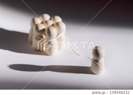 Wooden figurines representing social dynamics, one isolated figure stands apart from a group of connected figures tied with string on a white background 139540232