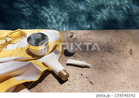 Skincare products including cream, serum, and sunscreen placed on a sunlit stone surface beside a swimming pool with clear blue water 139540242