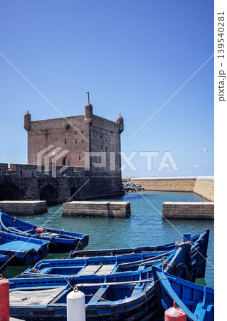 Blue fishing boats are moored in a tranquil harbor, with a historic stone tower standing majestically against a clear blue sky, creating a picturesque coastal scene 139540281
