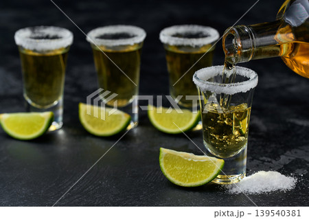 pouring golden tequila into a glass with lime slices and salt crystals on a dark table, selective focus. 139540381