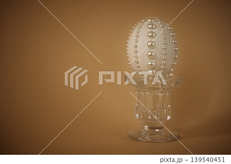 Decorative white egg adorned with pearl-like beads displayed on a clear glass pedestal against a warm brown background, ideal for festive or seasonal decor Decorative white egg adorned with pearl-like beads displayed on a clear glass pedestal against a warm brown background, ideal for festive or seasonal decor 139540451