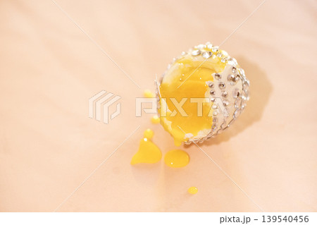 Decorative egg with silver embellishments cracked open, revealing bright yellow yolk and spilling liquid on a soft pink surface Decorative egg with silver embellishments cracked open, revealing bright yellow yolk and spilling liquid on a soft pink surface 139540456