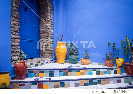 Colorful ceramic pots and plants on tiled steps leading to a blue door in a vibrant courtyard, showcasing traditional Moroccan architecture and design elements Colorful ceramic pots and plants on tiled steps leading to a blue door in a vibrant courtyard, showcasing traditional Moroccan architecture and design elements 139540465