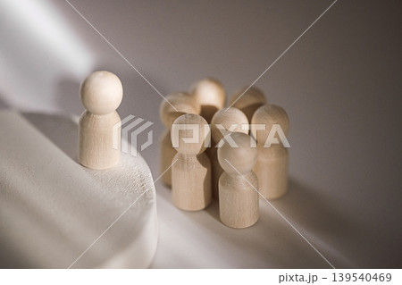Wooden figurines representing a group dynamic, one figure stands apart on a circular base while others are clustered together in a soft light setting Wooden figurines representing a group dynamic, one figure stands apart on a circular base while others are clustered together in a soft light setting 139540469