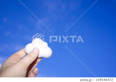 Hand holding a white cloud-shaped object against a clear blue sky, showcasing a whimsical and imaginative concept of clouds and weather Hand holding a white cloud-shaped object against a clear blue sky, showcasing a whimsical and imaginative concept of clouds and weather 139540472