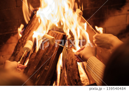Hands holding skewers with marshmallows roasting over a warm fire in a cozy setting, glowing flames and logs creating a festive atmosphere for gatherings 139540484