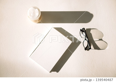 Minimalist composition featuring a blank book, stylish eyeglasses, and a cup of coffee, casting soft shadows on a light surface, creating a serene reading atmosphere Minimalist composition featuring a blank book, stylish eyeglasses, and a cup of coffee, casting soft shadows on a light surface, creating a serene reading atmosphere 139540487