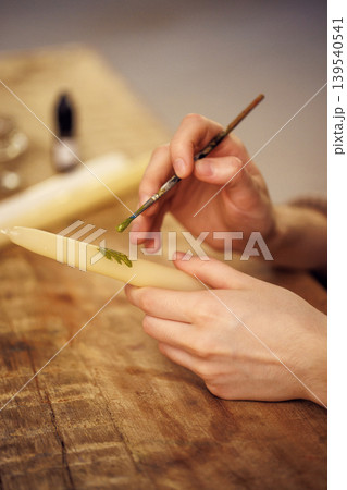 Hands of a creative individual painting intricate green designs on a white candle, showcasing artistic craftsmanship and attention to detail in a cozy workspace environment Hands of a creative individual painting intricate green designs on a white candle, showcasing artistic craftsmanship and attention to detail in a cozy workspace environment 139540541