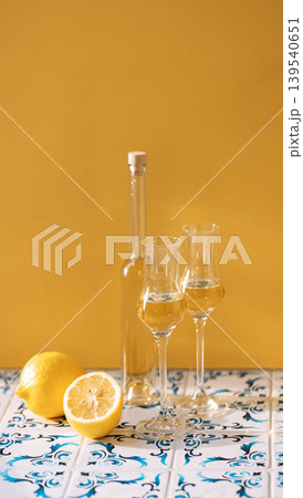 Filled grappa glasses and a bottle on the table with patterned tiles. Shot glasses with alcoholic drink and lemons. Minimalistic composition with glasses of limoncello. Filled grappa glasses and a bottle on the table with patterned tiles. Shot glasses with alcoholic drink and lemons. Minimalistic composition with glasses of limoncello. 139540651