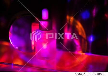Glass dropper bottle with a silver cap illuminated by colorful lights, creating a vibrant display of pink, purple, and blue reflections on a reflective surface 139540882