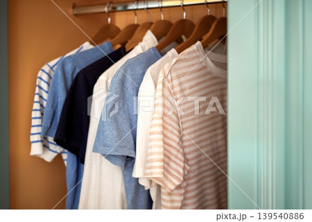 Collection of casual t-shirts in various colors and patterns hanging on wooden hangers inside a closet with a light blue door frame Collection of casual t-shirts in various colors and patterns hanging on wooden hangers inside a closet with a light blue door frame 139540886