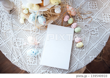 Easter-themed table setting with decorative pastel eggs and a blank notebook on a lace tablecloth, surrounded by festive decorations and a cake stand 139540910