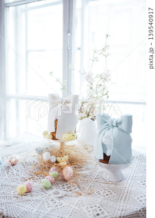 Decorative Easter cakes with white and blue bows displayed on white pedestals, surrounded by colorful eggs and spring flowers on a lace tablecloth near a bright window Decorative Easter cakes with white and blue bows displayed on white pedestals, surrounded by colorful eggs and spring flowers on a lace tablecloth near a bright window 139540917
